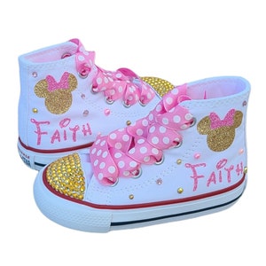 May include: White high-top trainers with pink and white polka dot ribbon laces. The trainers are decorated with gold glitter Mickey Mouse ears and the name "Faith" in pink glitter.