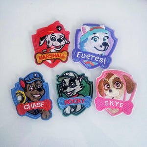 May include: Five embroidered patches featuring characters from a popular animated series. Each patch has a shield shape with a character's face and name. Colors include red, blue, green, purple, and pink. The names are Marshall, Everest, Chase, Rocky, and Skye.