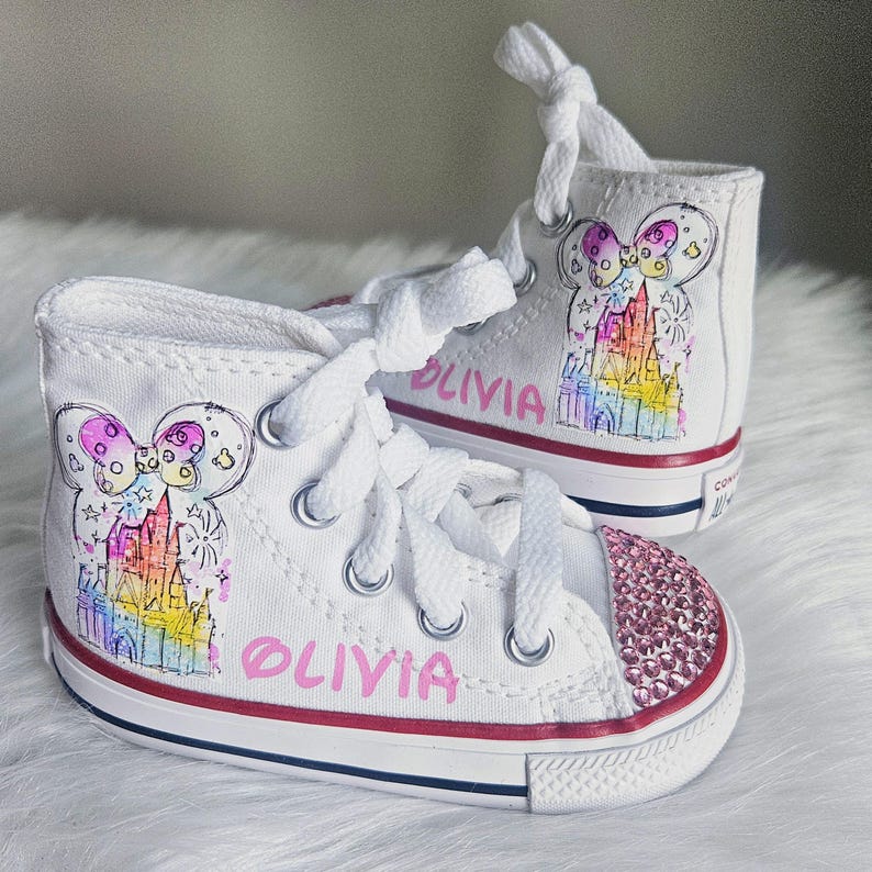 May include: White high-top trainers with a pink jeweled toe cap. Each shoe features a colourful castle design and the name "OLIVIA" in pink. The trainers have white laces and a red stripe along the sole.