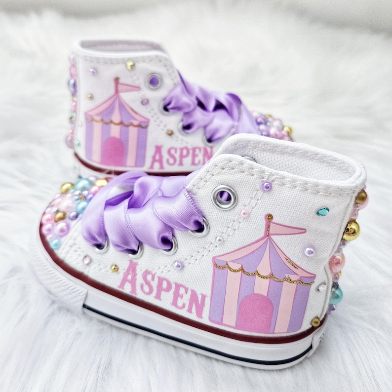 May include: White high-top sneakers with a pink and purple circus tent design and the name "Aspen" written on the side. The shoes are decorated with iridescent beads and a purple satin ribbon.