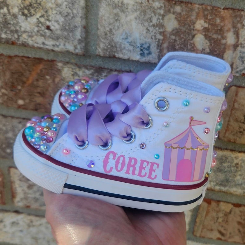 May include: White high-top sneakers with purple satin laces and a pink and purple circus tent design. The sneakers are decorated with iridescent rhinestones and the name "Coree" is written in pink on the side.