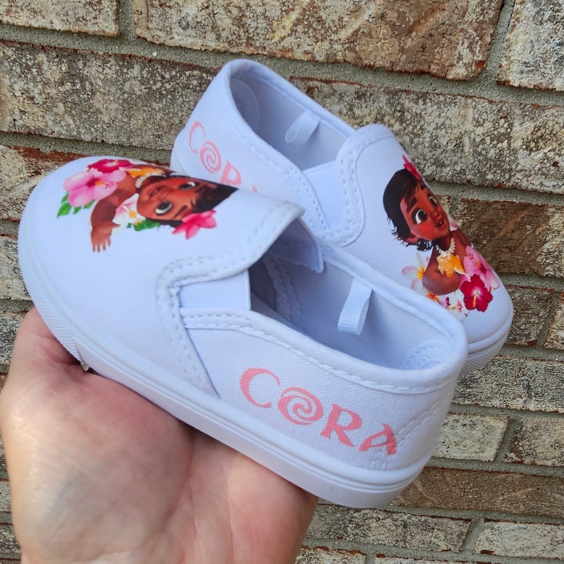 May include: White canvas slip-on shoes with a pink swirl design and the name "CORA" printed on the side. The shoes feature a cartoon image of Moana from the Disney movie Moana.