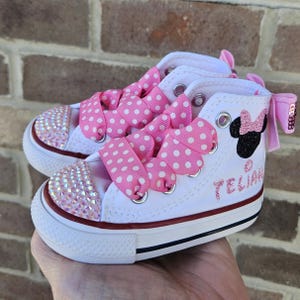 Puede incluir: Zapatillas Converse blancas de ca&ntilde;a alta con cordones de cinta rosa con lunares y un dise&ntilde;o de Minnie Mouse negro con purpurina. Las zapatillas est&aacute;n decoradas con pedrer&iacute;a iridiscente y el nombre "Telian" est&aacute; escrito en el lateral.