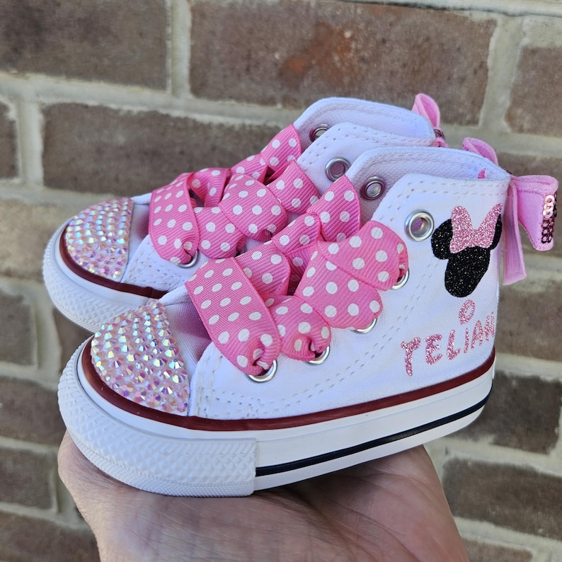 Puede incluir: Zapatillas Converse blancas de ca&ntilde;a alta con cordones de cinta rosa con lunares y un dise&ntilde;o de Minnie Mouse negro con purpurina. Las zapatillas est&aacute;n decoradas con pedrer&iacute;a iridiscente y el nombre "Telian" est&aacute; escrito en el lateral.