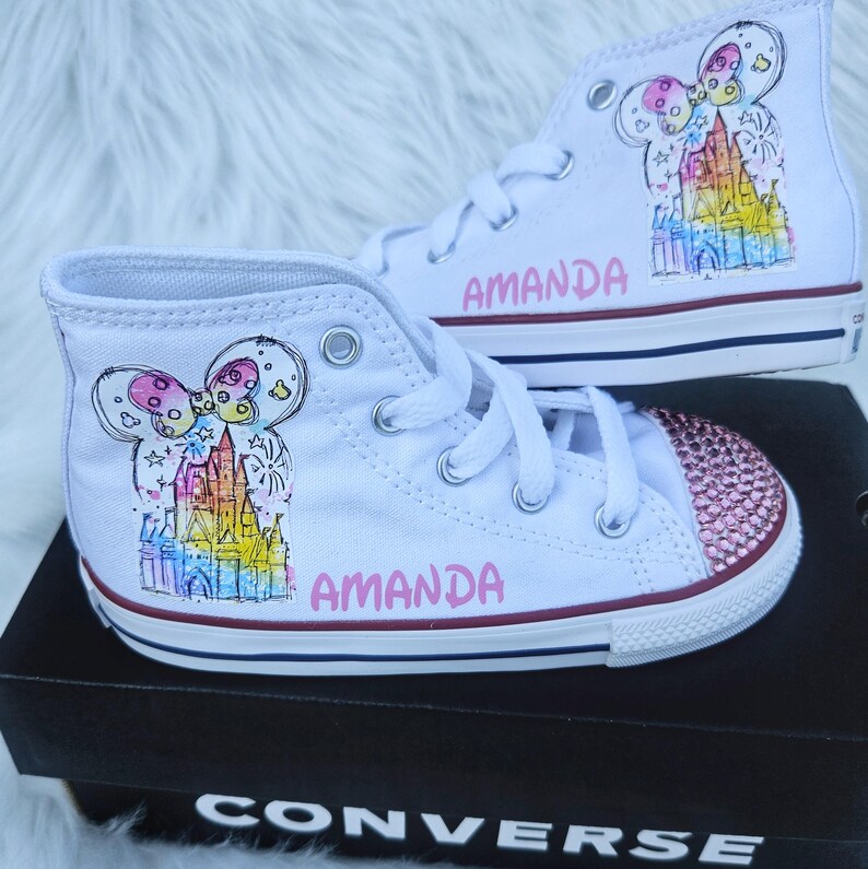 May include: White high-top Converse trainers with custom artwork. The trainers feature a colourful castle design and the name "AMANDA" in pink. The toe caps are embellished with pink rhinestones. The shoes are displayed on a black Converse box.