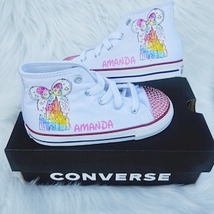 May include: White high-top trainers with custom artwork. The trainers feature a colourful castle design, a pink gem-studded toe cap, and the name "AMANDA" in pink lettering. The shoes are displayed on a black Converse box.