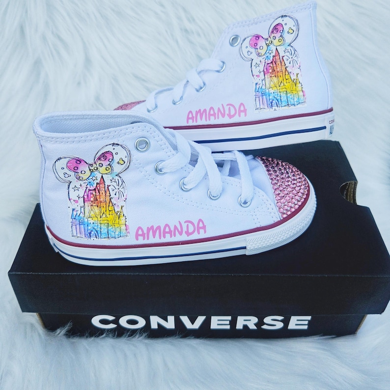 May include: White high-top trainers with custom artwork. The trainers feature a colourful castle design, a pink gem-studded toe cap, and the name "AMANDA" in pink lettering. The shoes are displayed on a black Converse box.