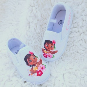 May include: A pair of white slip-on canvas shoes with a printed image of Moana from the Disney movie Moana. The shoes have pink flowers and a pink inscription that says "Cori" on one shoe and "5/6" on the other.