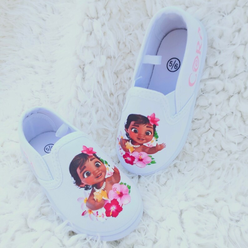 May include: A pair of white slip-on canvas shoes with a printed image of Moana from the Disney movie Moana. The shoes have pink flowers and a pink inscription that says "Cori" on one shoe and "5/6" on the other.