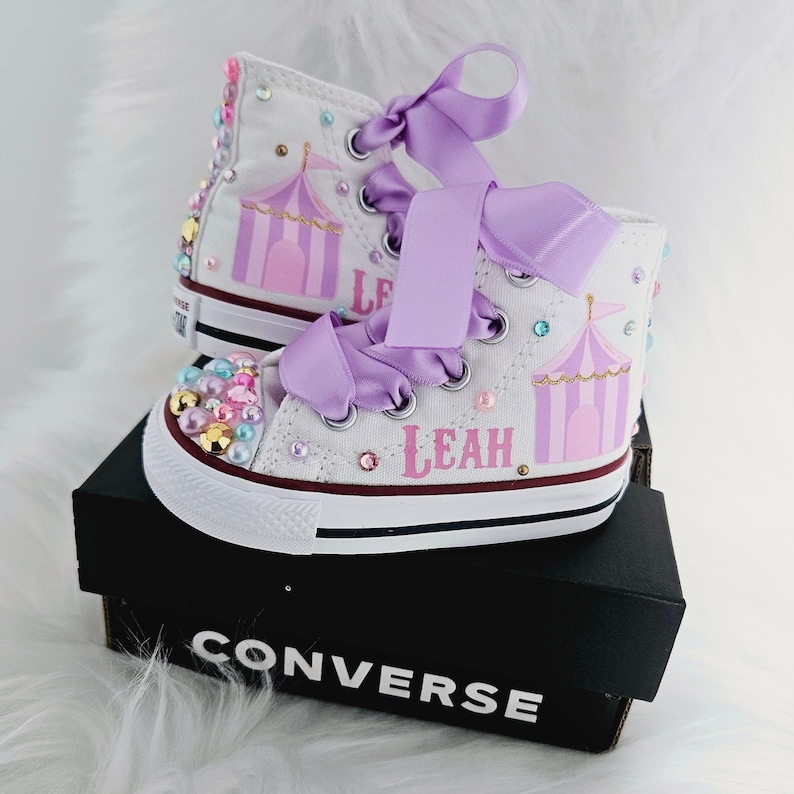 May include: White high-top Converse sneakers with a pink and gold circus tent design, purple satin laces, and the name "Leah" written in pink. The shoes are decorated with colorful rhinestones.