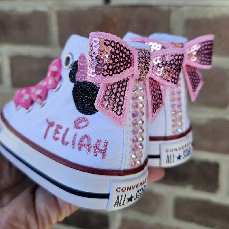Puede incluir: Zapatillas Converse blancas de ca&ntilde;a alta con detalles de Minnie Mouse en rosa y negro, un lazo rosa con lentejuelas y el nombre "Teliah" escrito en purpurina rosa.
