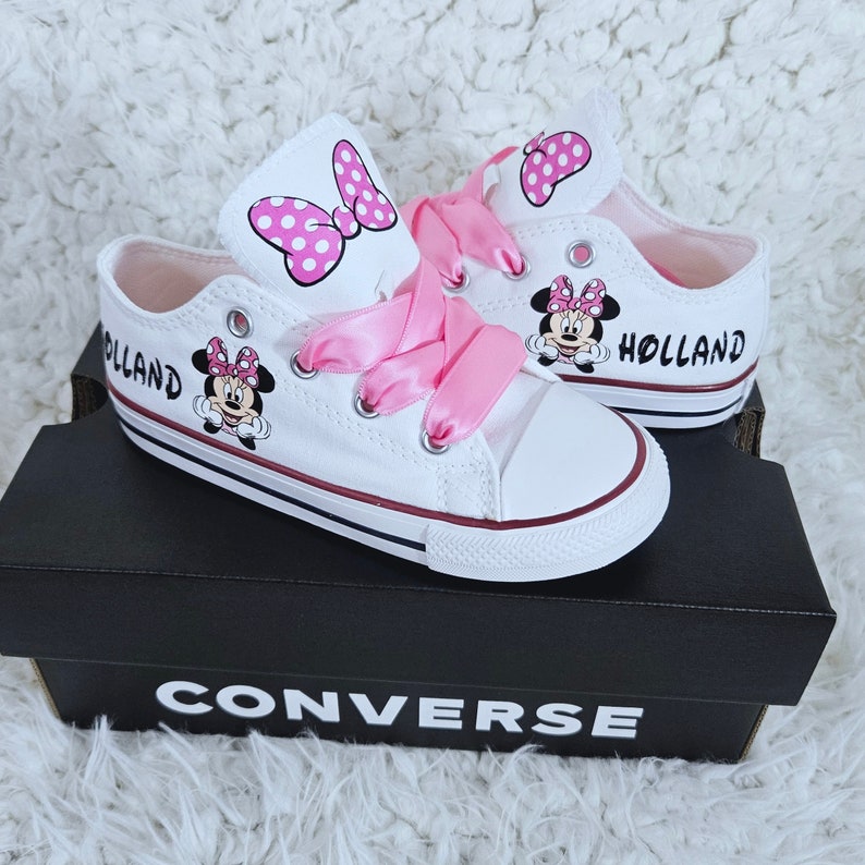 Pode incluir: T&ecirc;nis Converse brancos com cadar&ccedil;os de cetim rosa e um design de Minnie Mouse. Os t&ecirc;nis t&ecirc;m um la&ccedil;o rosa e bolinhas no lado e o nome "HOLLAND" escrito no lado.