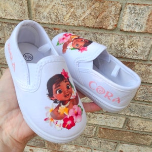 May include: White slip-on canvas shoes with a pink floral design and a cartoon image of Moana from Disney's Moana. The shoes are personalized with the name "Cora" in pink.