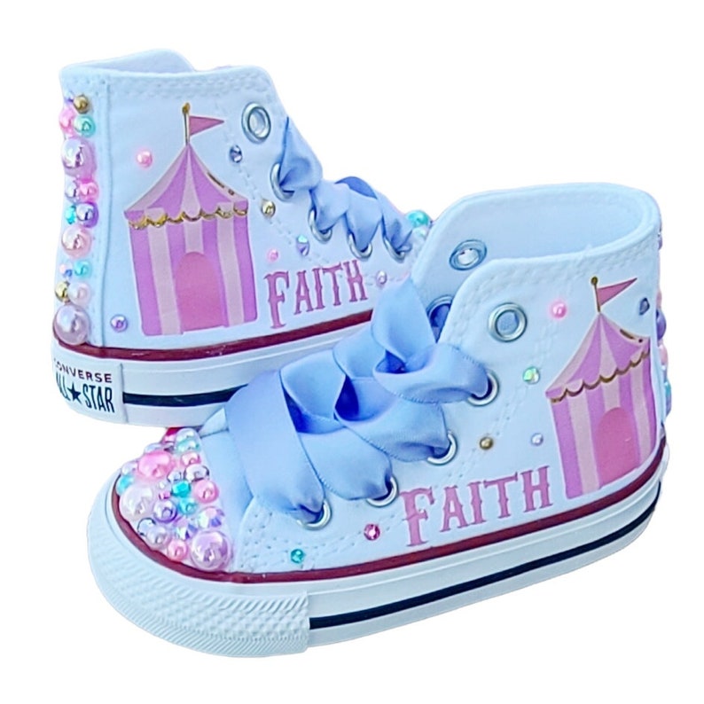 May include: Light blue high-top sneakers with a pink and white circus tent design and the name "Faith" written on them. The sneakers are decorated with iridescent gems.