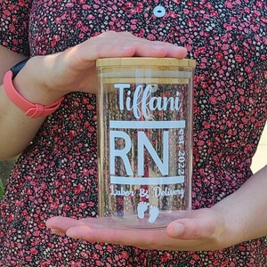 May include: A clear glass jar with a bamboo lid. The jar has a white label with the text "Tiffany RN est. 2022 Labor & Delivery" and two white footprints.