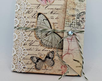 Puede incluir: Un diario hecho a mano con una estética vintage. La portada presenta ilustraciones de mariposas en rosa y negro, superpuestas sobre papel envejecido con escritura. Un ribete de encaje y cuerda con un acento de gema añaden detalles.