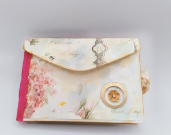 Puede incluir: Un diario de estilo vintage con un diseño floral y un acento de ojo de cerradura dorado. El diario tiene una cinta rosa y un círculo dorado con un diseño de flor. El texto "What Joy" está escrito en la portada.