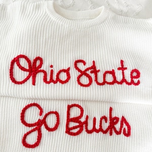 Könnte beinhalten: Zwei weiße Pullover mit rotem gesticktem Text. Der obere Pullover lautet "Ohio State" und der untere Pullover lautet "Go Bucks". Die Pullover sind aus einem Strickmaterial gefertigt.
