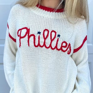 Peut inclure: Pull en tricot crème avec des accents rouges et le mot "Phillies" brodé en rouge sur la poitrine. Le pull a une bordure rouge sur l'encolure et les manches, et des rayures rouges sur les manches.