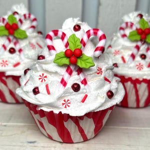 May include: Three decorative cupcakes with white frosting, red and white striped wrappers, and candy cane accents. Each cupcake is topped with candy canes, holly leaves, and red berries, with red sprinkles and snowflake decorations.