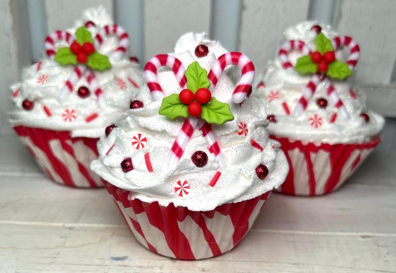 May include: Three decorative cupcakes with white frosting, red and white striped wrappers, and candy cane accents. Each cupcake is topped with candy canes, holly leaves, and red berries, with red sprinkles and snowflake decorations.