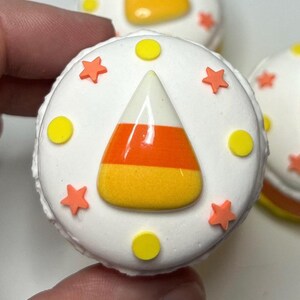 Puede incluir: Primer plano de un macaron redondo blanco con tem&aacute;tica de Halloween decorado con un dise&ntilde;o de caramelo de ma&iacute;z. El caramelo de ma&iacute;z es naranja, amarillo y blanco. El macaron est&aacute; decorado con chispas de estrellas naranjas y c&iacute;rculos amarillos.