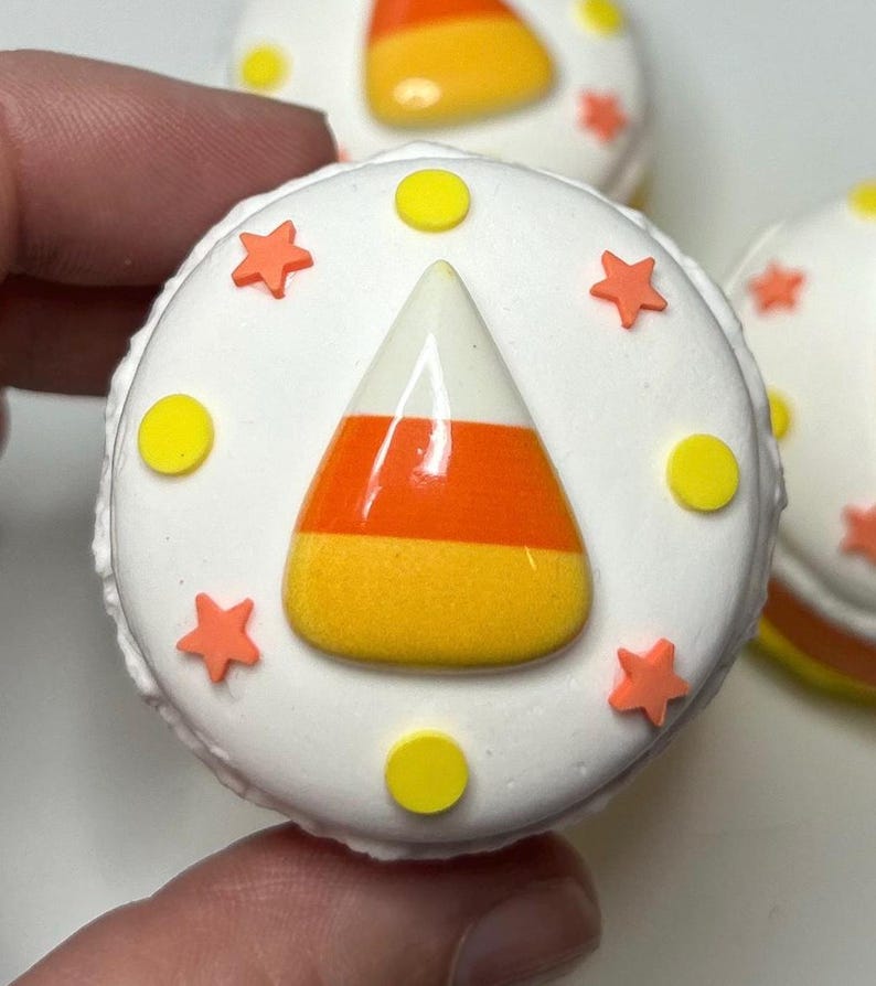 Puede incluir: Primer plano de un macaron redondo blanco con tem&aacute;tica de Halloween decorado con un dise&ntilde;o de caramelo de ma&iacute;z. El caramelo de ma&iacute;z es naranja, amarillo y blanco. El macaron est&aacute; decorado con chispas de estrellas naranjas y c&iacute;rculos amarillos.