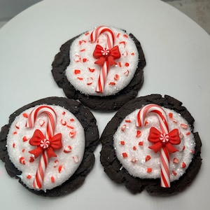 May include: Three chocolate cookies decorated with white frosting and red and white candy canes. The cookies are on a white surface.