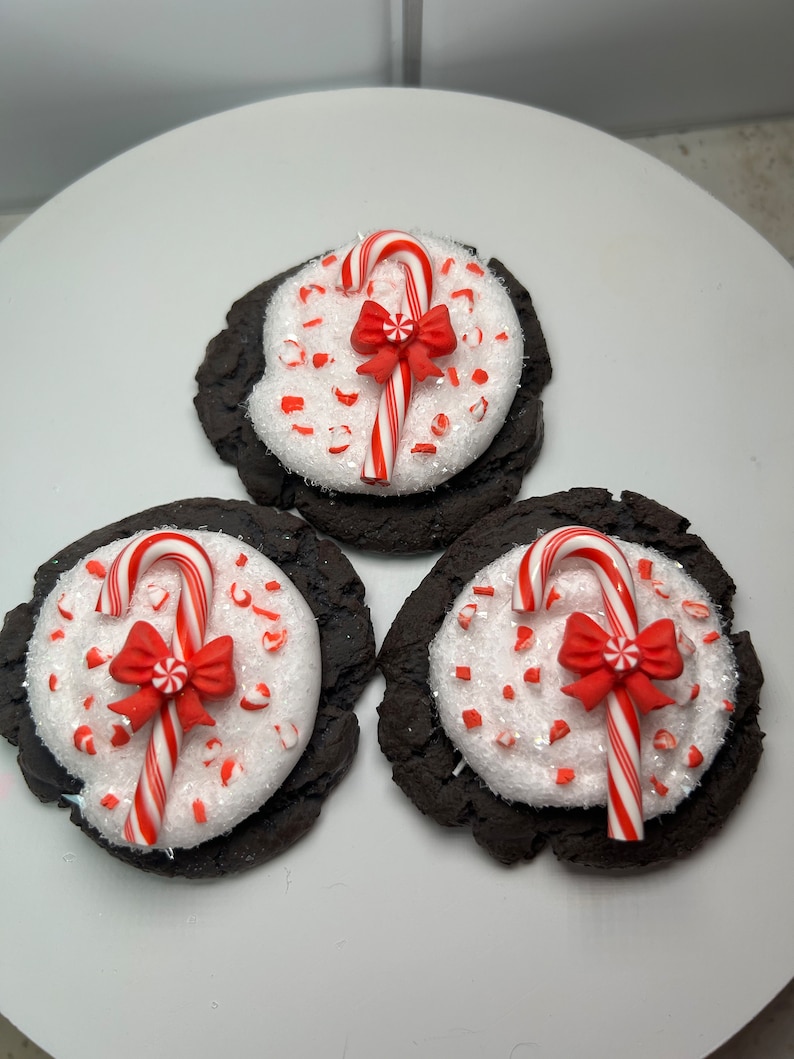 May include: Three chocolate cookies decorated with white frosting and red and white candy canes. The cookies are on a white surface.