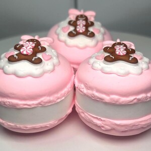 May include: Three pink macaron-shaped objects with white frosting and gingerbread man decorations. Each macaron has a pink bow on top and a white filling. The gingerbread men have pink and white candy accents.