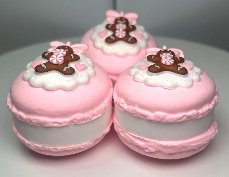 May include: Three pink macaron-shaped objects with white frosting and gingerbread man decorations. Each macaron has a pink bow on top and a white filling. The gingerbread men have pink and white candy accents.