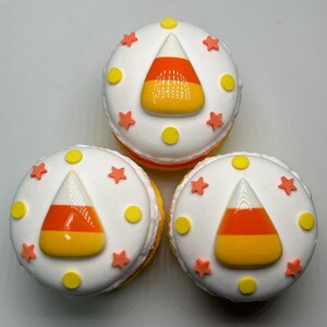 Puede incluir: Tres macarons blancos con decoraciones de caramelos de ma&iacute;z naranja, amarillo y blanco. Cada macaron est&aacute; decorado con estrellas naranjas y puntos amarillos. Los macarons son un dulce festivo.