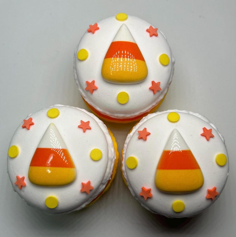 Puede incluir: Tres macarons blancos con decoraciones de caramelos de ma&iacute;z naranja, amarillo y blanco. Cada macaron est&aacute; decorado con estrellas naranjas y puntos amarillos. Los macarons son un dulce festivo.