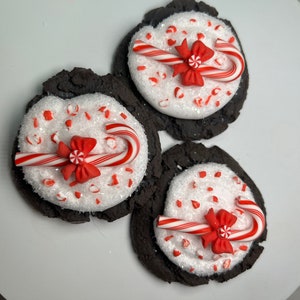 May include: Three chocolate cookies decorated with white frosting, red and white candy canes, and red bows. The cookies are on a white plate.