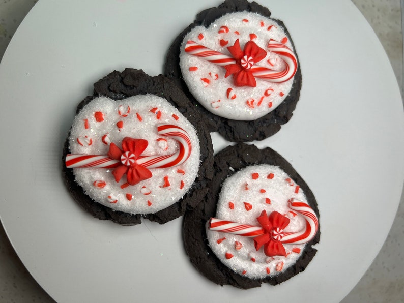 May include: Three chocolate cookies decorated with white frosting, red and white candy canes, and red bows. The cookies are on a white plate.