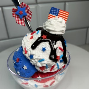 May include: A red, white, and blue ice cream sundae with chocolate sauce, sprinkles, and a miniature American flag on top. The sundae is in a clear glass bowl.