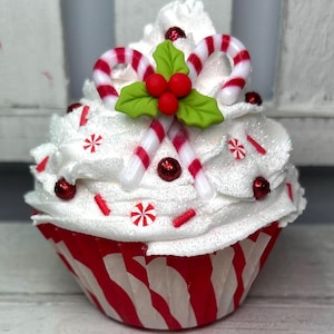 May include: A festive cupcake with white frosting, red and white candy canes, and red sprinkles. The cupcake is in a red and white striped paper cup, decorated with red glitter and holly leaves. A holiday-themed dessert.