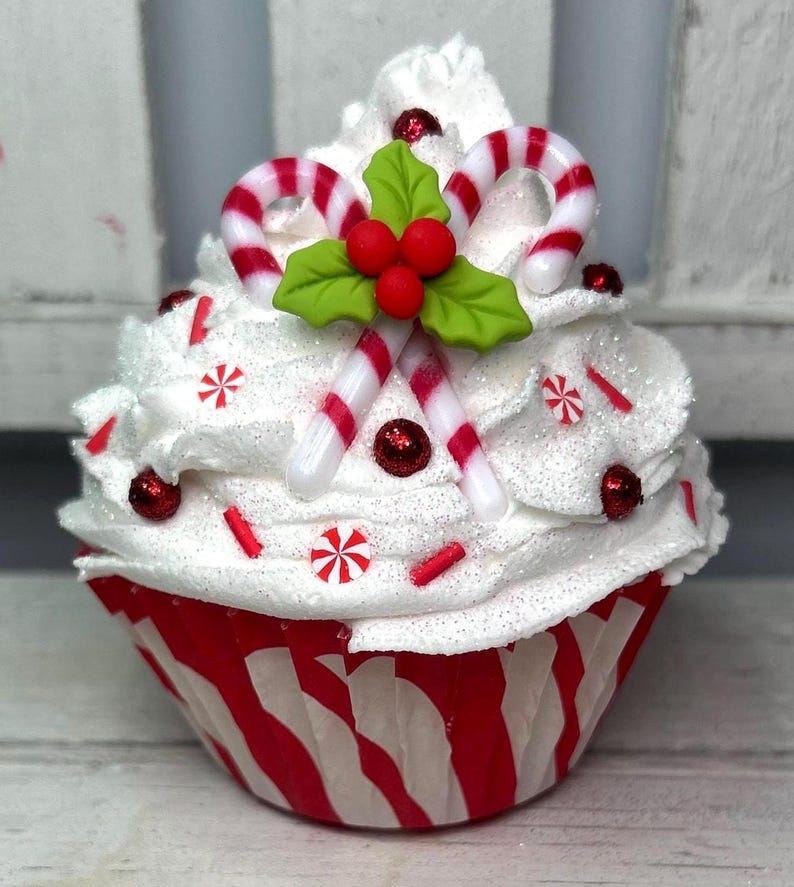 May include: A festive cupcake with white frosting, red and white candy canes, and red sprinkles. The cupcake is in a red and white striped paper cup, decorated with red glitter and holly leaves. A holiday-themed dessert.