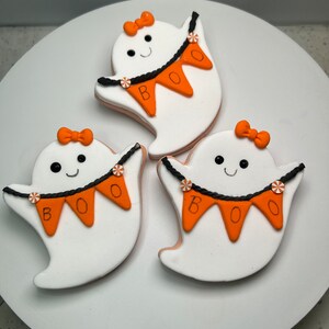 Pode incluir: Tr&ecirc;s biscoitos brancos em forma de fantasma decorados com glac&ecirc; laranja e preto. Cada biscoito tem um la&ccedil;o preto, um rosto sorridente e uma faixa que diz "BOO".