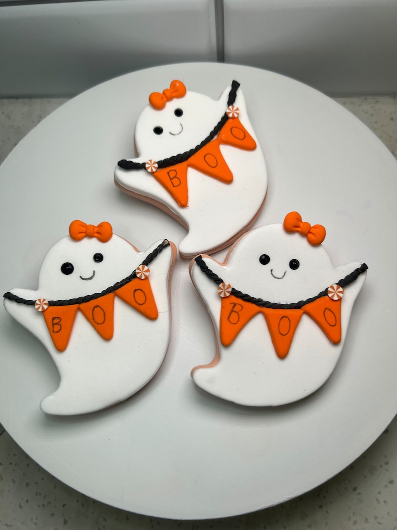 Pode incluir: Tr&ecirc;s biscoitos brancos em forma de fantasma decorados com glac&ecirc; laranja e preto. Cada biscoito tem um la&ccedil;o preto, um rosto sorridente e uma faixa que diz "BOO".