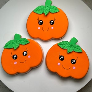 Pode incluir: Tr&ecirc;s biscoitos em forma de ab&oacute;bora laranja decorados com folhas verdes, olhos pretos e bochechas cor-de-rosa. Cada biscoito tem um rosto sorridente.
