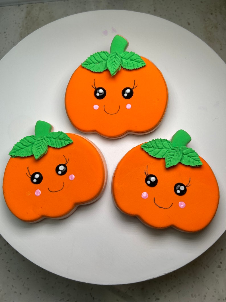 Pode incluir: Tr&ecirc;s biscoitos em forma de ab&oacute;bora laranja decorados com folhas verdes, olhos pretos e bochechas cor-de-rosa. Cada biscoito tem um rosto sorridente.