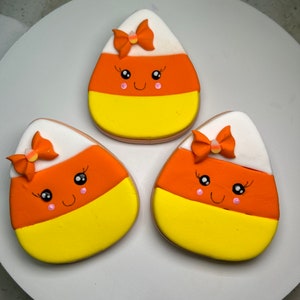 Pode incluir: Tr&ecirc;s biscoitos de a&ccedil;&uacute;car decorados em forma de milho doce. Cada biscoito tem um desenho de glac&ecirc; branco, laranja e amarelo com um la&ccedil;o rosa e branco no topo. Os biscoitos t&ecirc;m um rosto de desenho animado bonito com bochechas cor-de-rosa e olhos pretos.