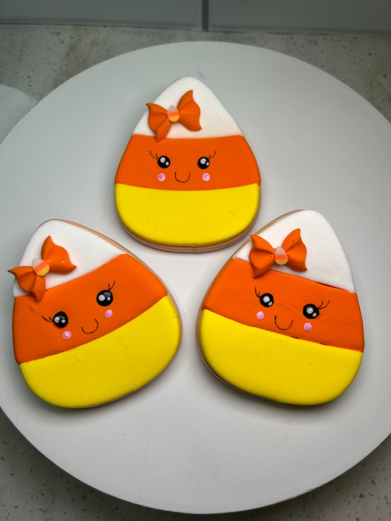 Pode incluir: Tr&ecirc;s biscoitos de a&ccedil;&uacute;car decorados em forma de milho doce. Cada biscoito tem um desenho de glac&ecirc; branco, laranja e amarelo com um la&ccedil;o rosa e branco no topo. Os biscoitos t&ecirc;m um rosto de desenho animado bonito com bochechas cor-de-rosa e olhos pretos.