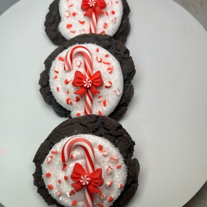 May include: Three chocolate cookies decorated with white frosting, red and white candy canes, and red bows. The cookies are arranged vertically on a white plate.