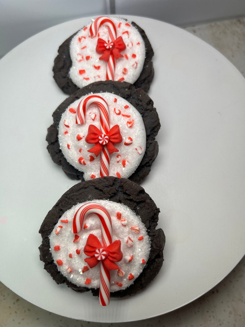 May include: Three chocolate cookies decorated with white frosting, red and white candy canes, and red bows. The cookies are arranged vertically on a white plate.
