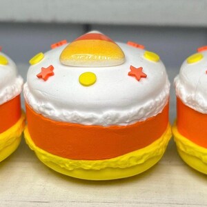 Puede incluir: Tres recipientes decorativos con forma de caramelo de ma&iacute;z. Cada uno tiene una parte superior blanca con detalles amarillos y naranjas, una parte media de color naranja brillante y una base amarilla. Las tapas est&aacute;n decoradas con formas de estrella y c&iacute;rculo.