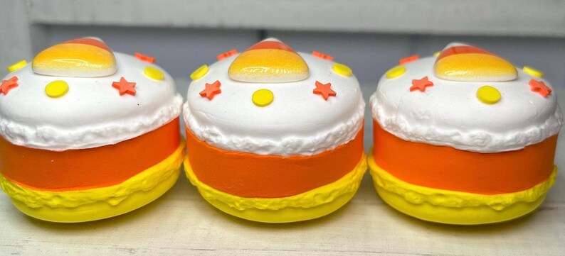 Puede incluir: Tres recipientes decorativos con forma de caramelo de ma&iacute;z. Cada uno tiene una parte superior blanca con detalles amarillos y naranjas, una parte media de color naranja brillante y una base amarilla. Las tapas est&aacute;n decoradas con formas de estrella y c&iacute;rculo.