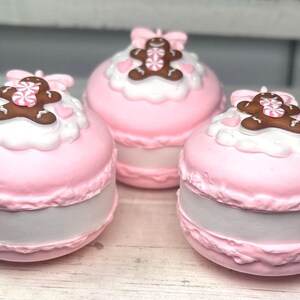 May include: Three pink, round decorative boxes resembling macarons. Each box has a white frosting layer topped with a gingerbread man and a pink bow. The boxes have a light gray base and are arranged on a white surface.