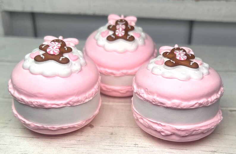 May include: Three pink, round decorative boxes resembling macarons. Each box has a white frosting layer topped with a gingerbread man and a pink bow. The boxes have a light gray base and are arranged on a white surface.